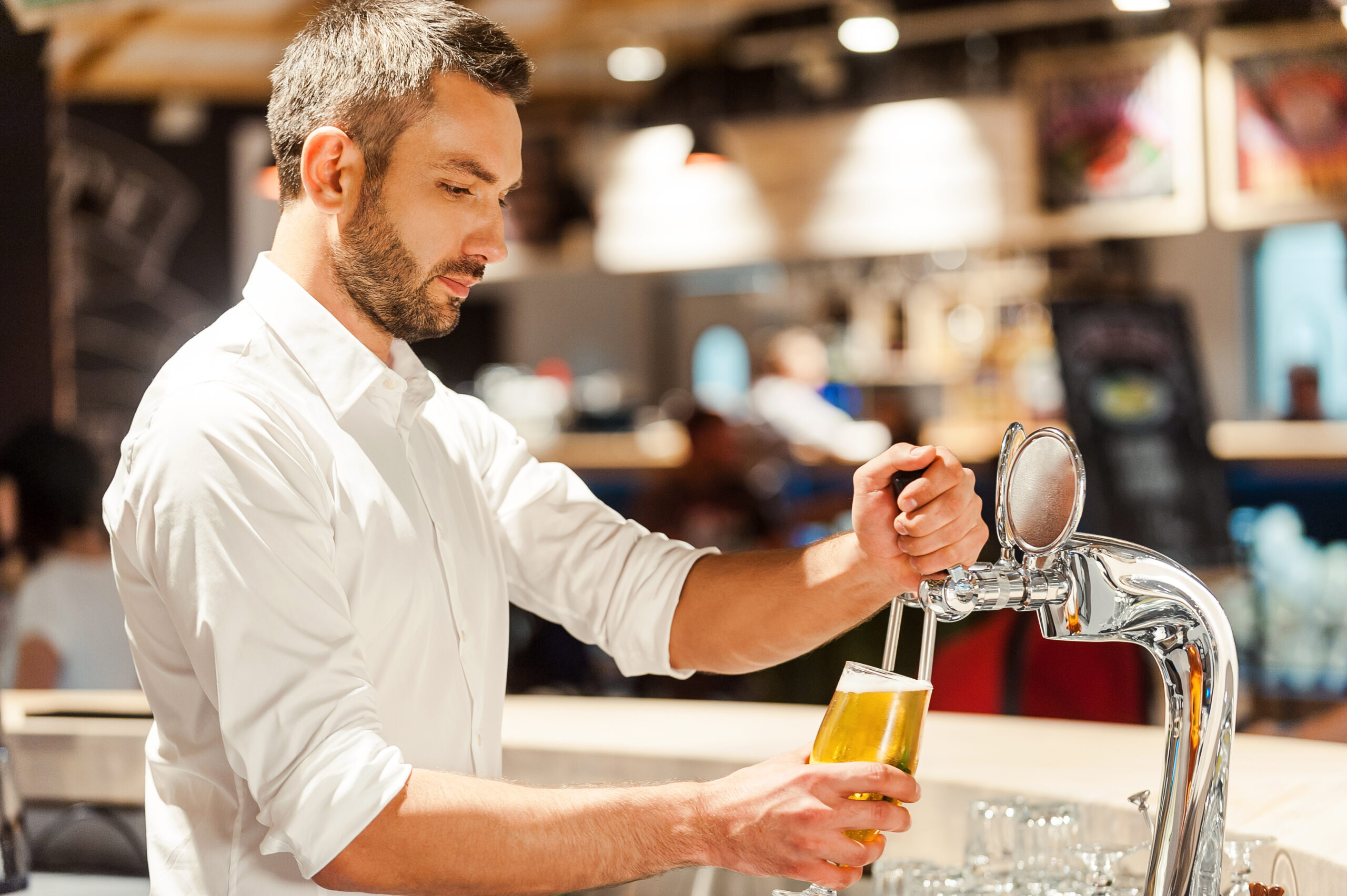 Pouring beer for client. Side view of young bartender pouring beer while standing at the bar counter